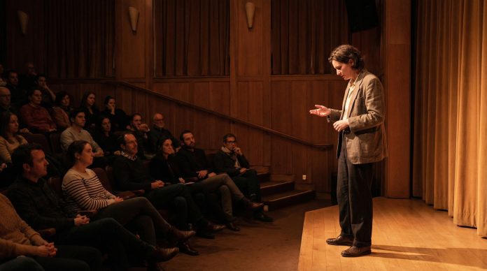 Thoughtful introverted speaker standing confidently on stage with soft spotlight and attentive audience